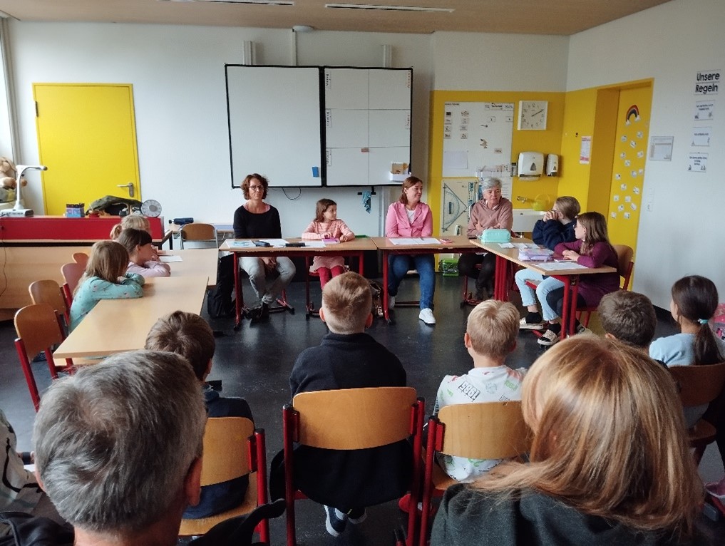 Kinder sitzen mit Tischen im Kreis neben Yvonne Magwas