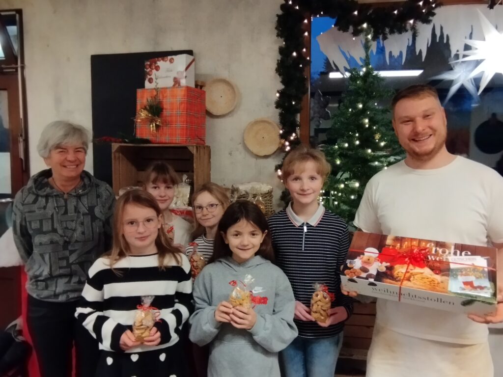 Bürgermeisterin Marion Dick mit dem Kindergemeinderat und Bäckermeister Steve Trützschler bei Übergabe der gesponserten Tombolapreise in der Bäckerfiliale. Die Kinder halten eine Kekstüte in der Hand, der Bäcker einen Weihnachtsstollen mit Gutscheinen.