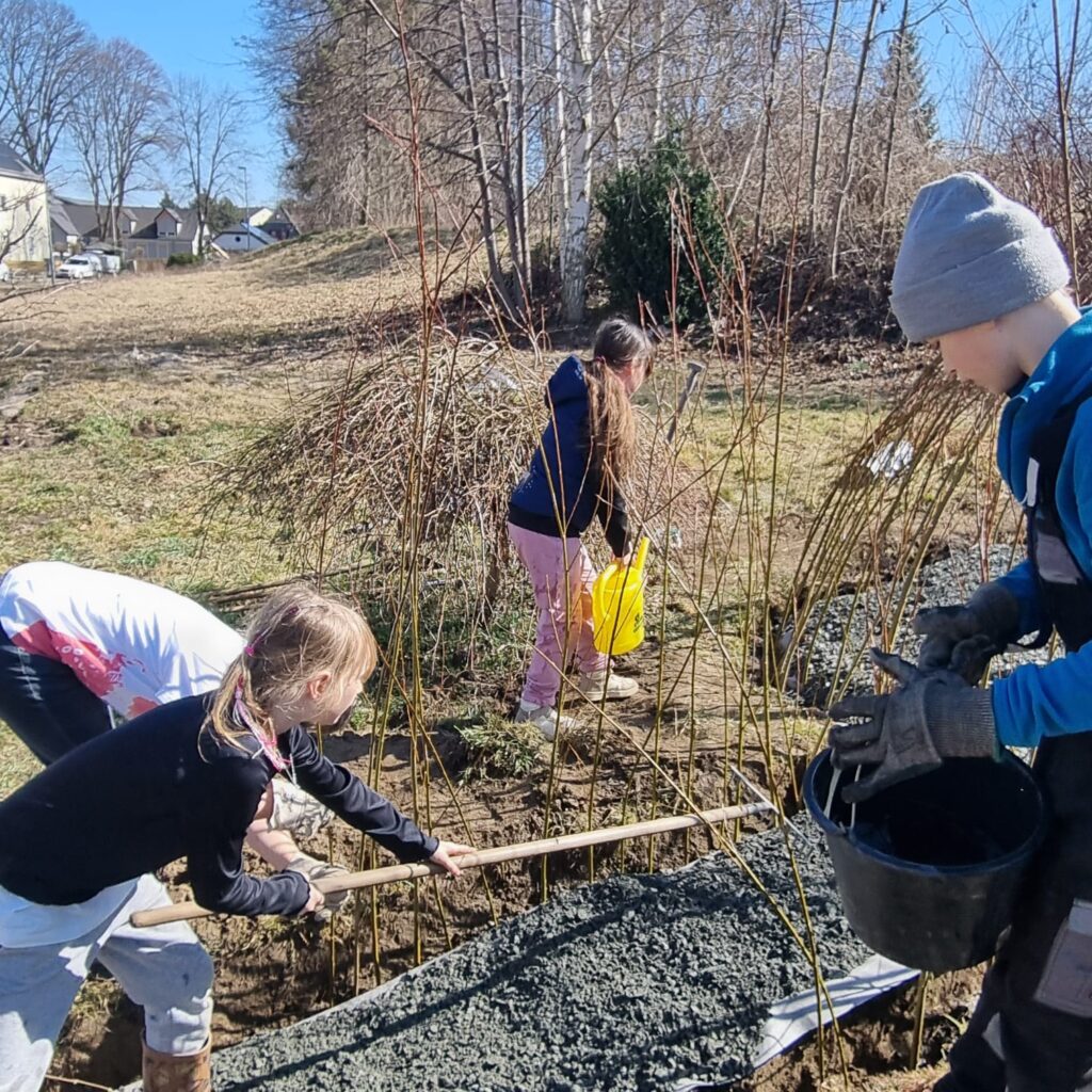 Auf dem Bild sind Kinder zu sehen, die an dem Weideniglu bauen (gießen und Splitt verteilen)